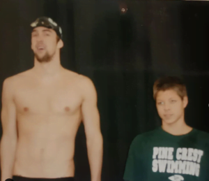 Um homem alto, sem camisa e com touca e óculos de natação ao lado de um menino mais baixo e com camiseta verde